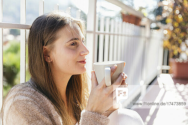 Junge Frau mit Kaffeetasse denkt nach  während sie auf dem Balkon sitzt