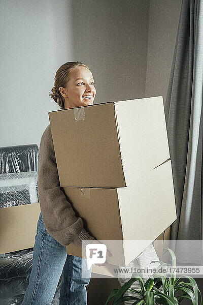 Smiling young woman carrying cardboard boxes in new apartment