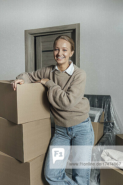 Smiling young woman leaning on stack of cartons at home
