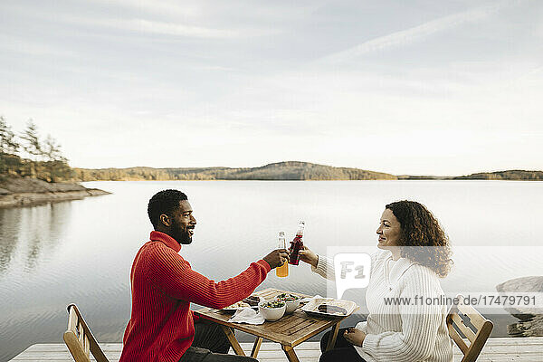 Lächelndes Paar stößt auf Flaschen an  während es am Wochenende auf einem Stuhl am See sitzt