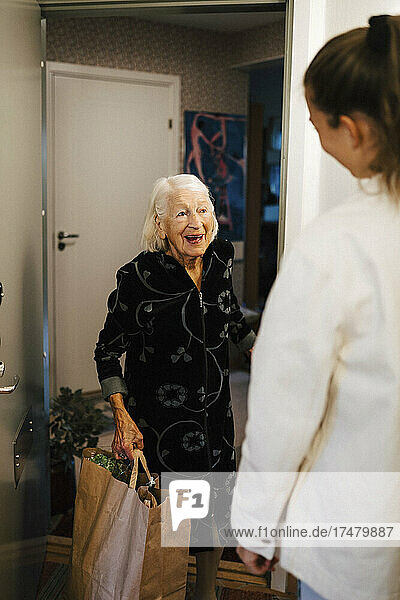 Glückliche ältere Frau mit Einkaufstasche  die eine Mitarbeiterin des Gesundheitswesens beim Betreten des Hauses beobachtet