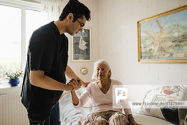 Male caregiver holding hand while giving support to senior woman sitting on bed at home