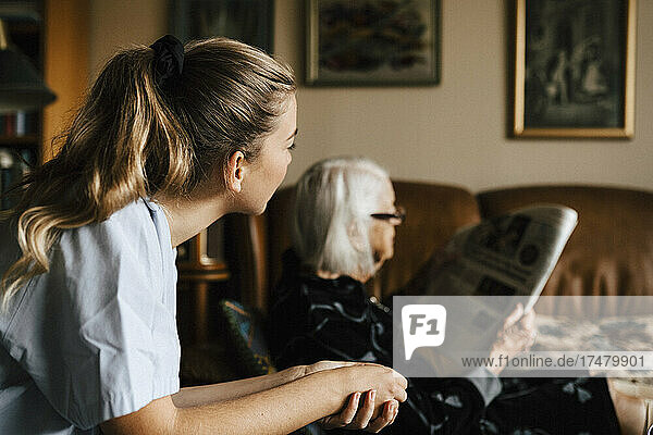 Weibliche Krankenschwester betrachtet eine ältere Frau  die im Wohnzimmer Zeitung liest