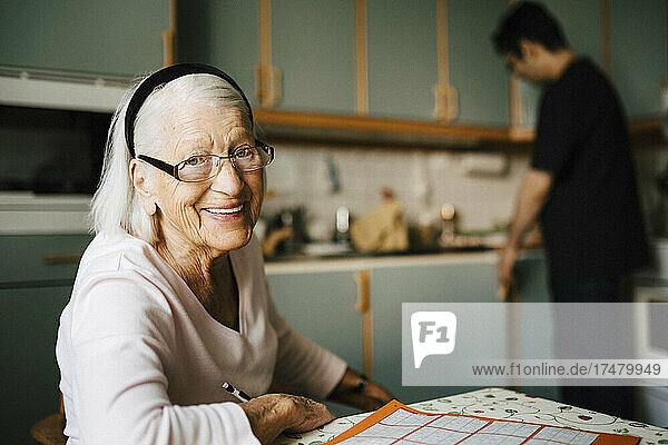 Porträt einer lächelnden älteren Frau  während im Hintergrund ein männlicher Pfleger zu Hause steht