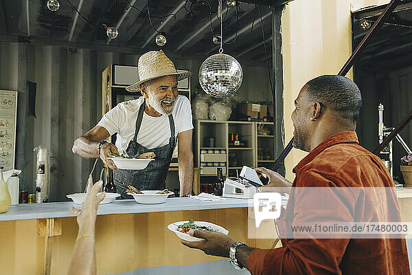 Smiling owner serving food while male customer doing contactless payment through smart phone