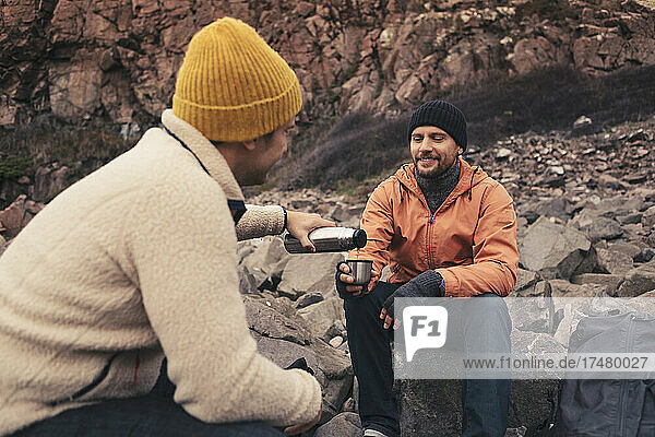 Man pouring drink for male friend while sitting on rock
