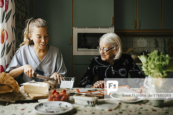 Weibliche Pflegekraft schneidet Brotlaib am Esstisch  während sie neben einer älteren Frau in der Küche sitzt