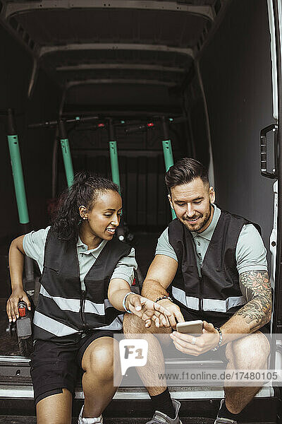 Lächelnde weibliche und männliche Mitarbeiter  die ihr Smartphone benutzen  während sie im Lieferwagen sitzen