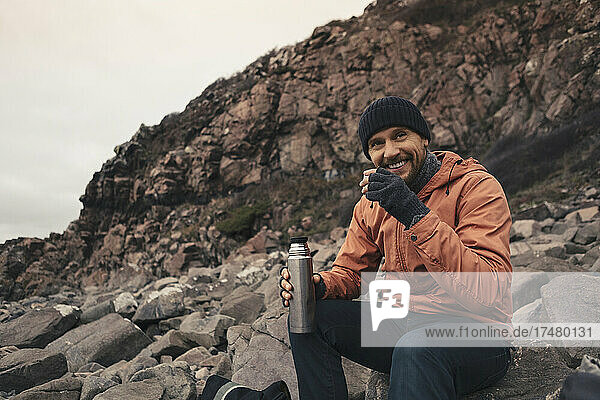 Lächelnder Mann  der auf einem Felsen am Strand sitzt und etwas trinkt