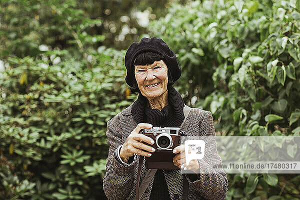 Porträt einer lächelnden älteren Frau mit Kamera vor Pflanzen im Park während des Urlaubs