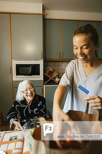 Glückliche ältere Frau  die eine Mitarbeiterin des Gesundheitswesens betrachtet  die in der Küche Lebensmittel aus der Tüte nimmt