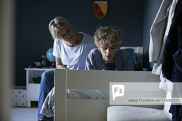 Mutter betrachtet ihren Sohn bei der Benutzung eines Smartphones auf dem Bett im Schlafzimmer