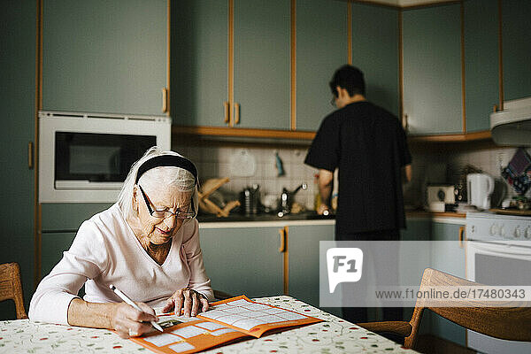 Elderly woman solving crossword puzzle in book while male nurse working at kitchen