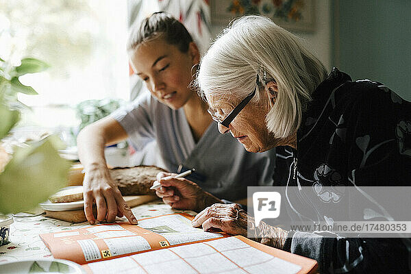 Female nurse solving crossword puzzle with senior woman while sitting in kitchen at home