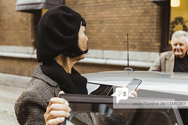 Ältere Frau mit Smartphone schaut Mann beim Einsteigen ins Auto in der Stadt an