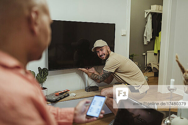 Lächelnder männlicher Techniker schaut auf einen reifen Mann  der ein Smartphone im Wohnzimmer benutzt