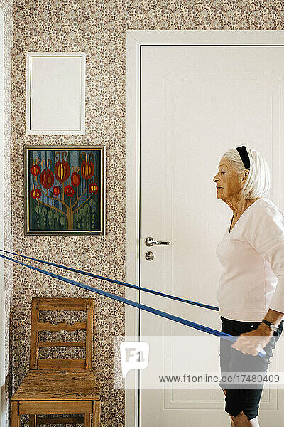 Seitenansicht einer älteren Frau,  die an einem Widerstandsband zieht,  während sie zu Hause trainiert