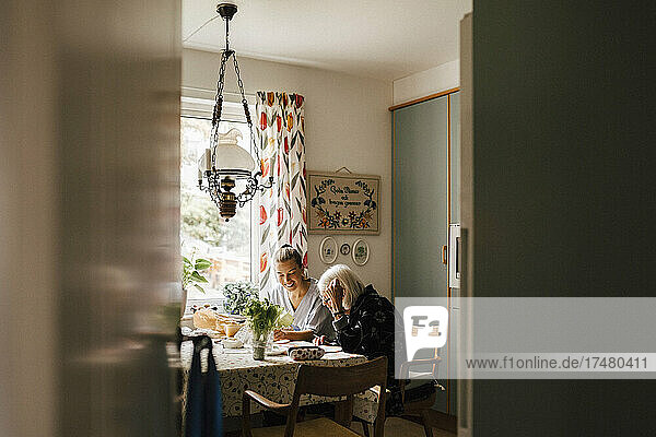 Ältere Frau  die in der Küche zu Hause bei einer weiblichen Pflegekraft sitzt und Zeitung liest