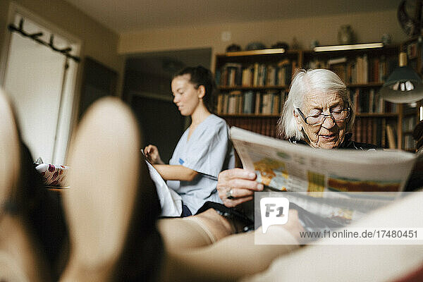 Ältere Frau liest Zeitung  während im Hintergrund eine Krankenschwester zu Hause ist