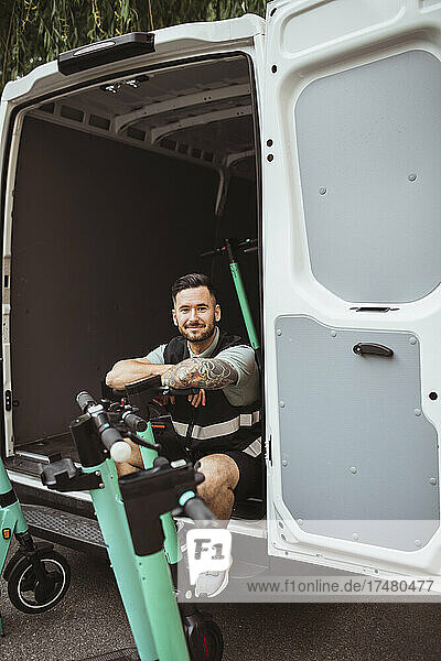 Porträt eines lächelnden männlichen Arbeiters  der in einem Lieferwagen sitzt und sich auf einen Elektroroller stützt