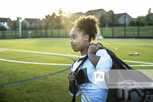 UK  Rear view of female soccer player (12-13) carrying bag with balls in field