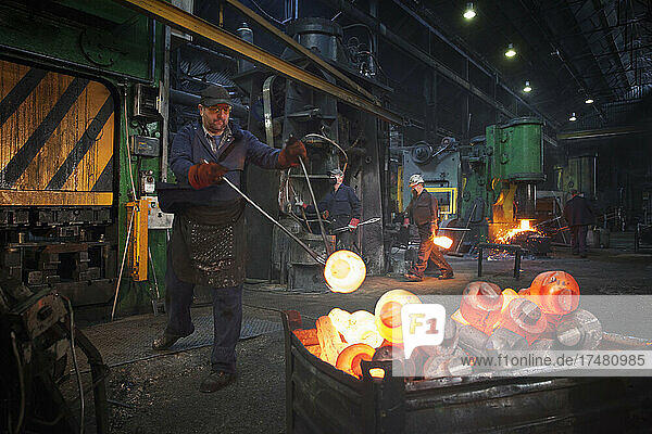 Forge worker swings finished forged component onto pallet as team works in background