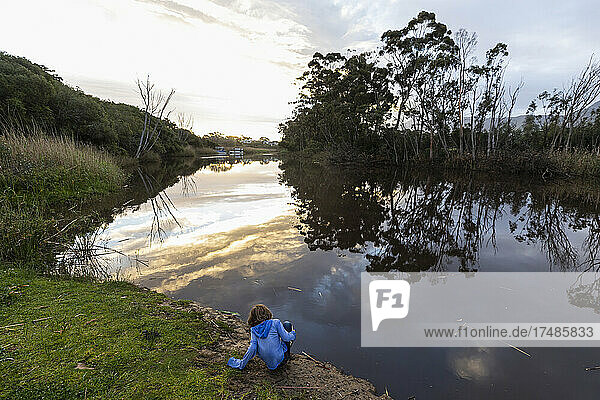 A young boy standing by a river at dusk  sky reflections in the flat calm water