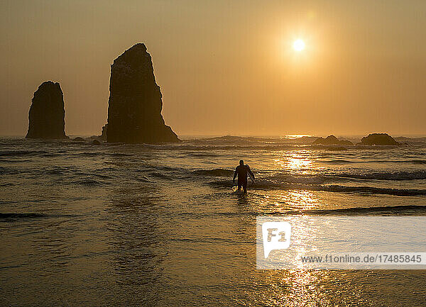 Surfer  der aus dem Meer steigt  während die Sonne hinter einem Felsen in den Wellen am Strand untergeht.