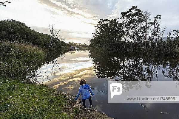 A young boy standing by a river at dusk  sky reflections in the flat calm water