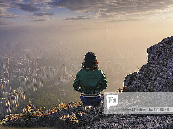 Frau sitzt auf einem Felsen in den Hügeln über Hongkong und blickt auf die Stadt hinunter.