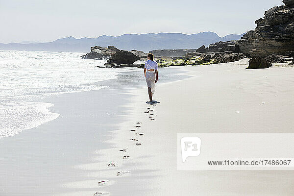 Jugendliches Mädchen  das allein an einem Sandstrand in Südafrika am Rande des Wassers spazieren geht  Rückansicht