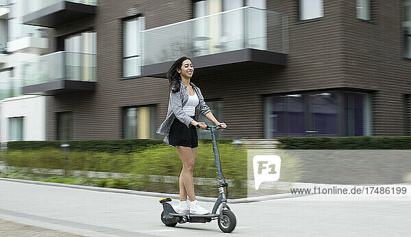 Fröhliche junge Frau fährt mit einem Elektroroller auf einem Bürgersteig