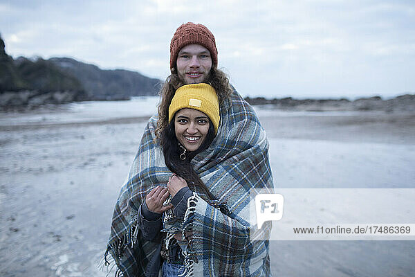 Portrait happy young couple wrapped in blanket on cold beach