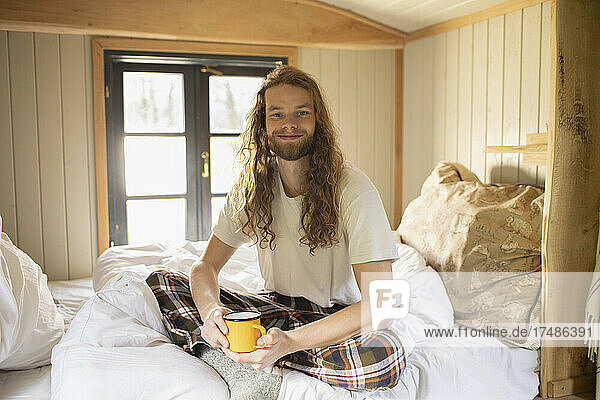 Porträt glücklicher junger Mann im Pyjama  der im Bett Kaffee trinkt
