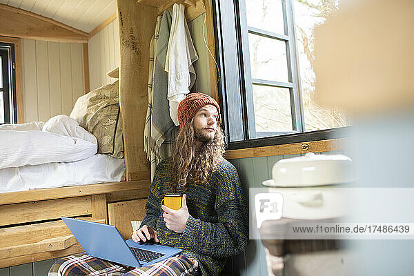 Junger Mann mit Kaffee und Laptop in einer kleinen Hütte