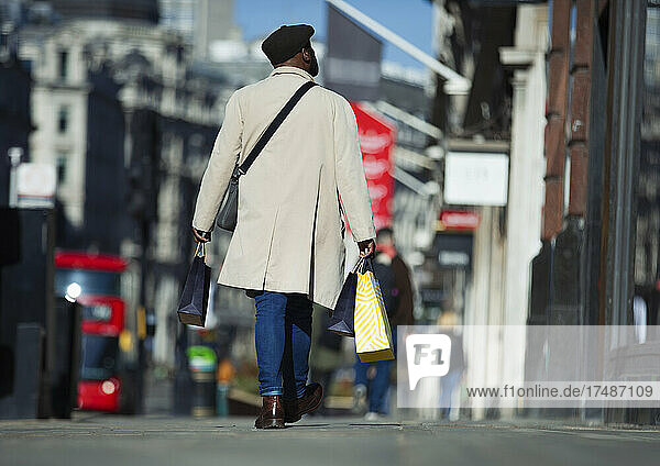 Mann im Trenchcoat trägt Einkaufstüten auf dem sonnigen Bürgersteig der Stadt