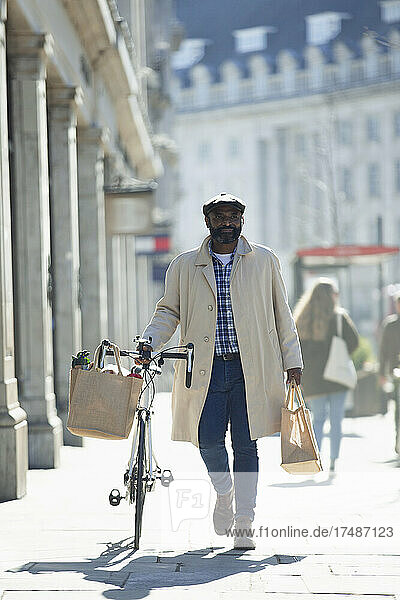 Man with bicycle and grocery bags walking on sunny sidewalk