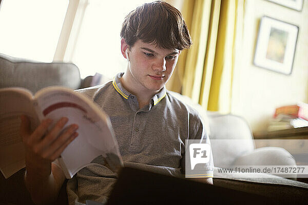 Focused teenage boy with textbook and laptop studying at home