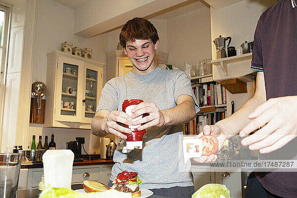 Glücklicher Teenager  der in der Küche Ketchup auf einen Hamburger drückt
