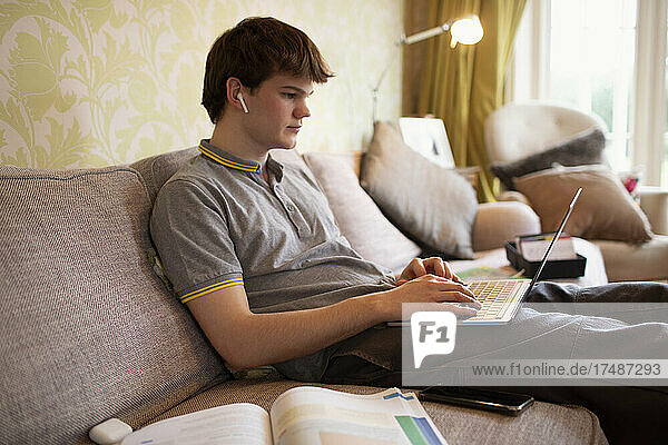 Teenage boy with earbuds using laptop on living room sofa