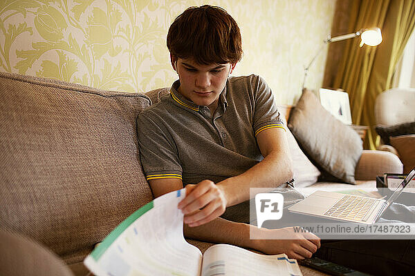 Jugendlicher mit Laptop und Lehrbuch beim Lernen zu Hause
