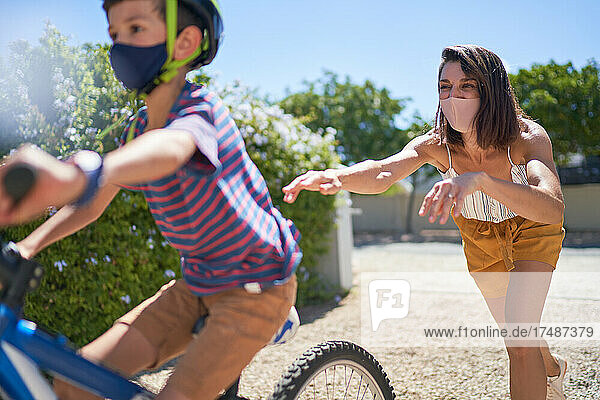 Mutter mit Gesichtsmaske schiebt Sohn auf Fahrrad in sonniger Einfahrt