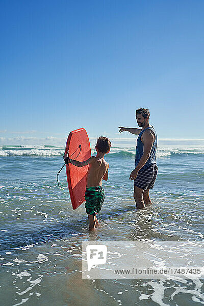 Vater und Sohn mit Bodyboard waten in der sonnigen Meeresbrandung