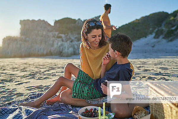 Glückliche Mutter und Sohn essen auf einer Picknickdecke am sonnigen Strand