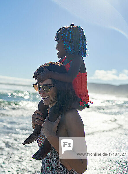 Glückliche Mutter trägt süße Tochter auf den Schultern am sonnigen Strand