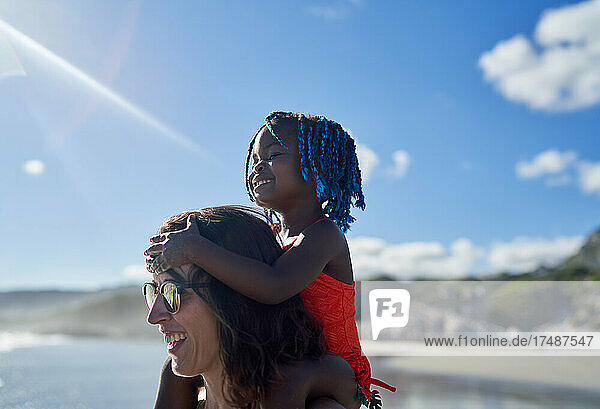 Glückliche Mutter trägt süße Tochter auf den Schultern am sonnigen Strand
