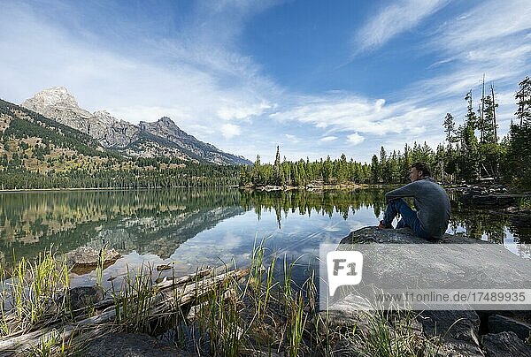 Junger Mann sitzt auf einem Stein an einem See  Spiegelung im Taggart Lake  Blick auf den Teton Range Gebirgszug  Gipfel Grand Teton und Teewinot Mountain  Grand Teton National Park  Wyoming  USA  Nordamerika