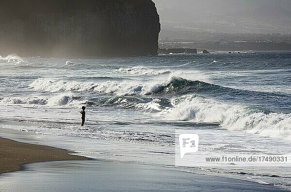 Eine Frau steht am Sandstrand von Praia de Santa Barbara bei stürmischen Meer mit hohen Wellen  Ribeira Grande  Insel Sao Miguel  Azoren  Portugal  Europa