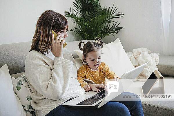 Mother with laptop talking on mobile phone by daughter at home