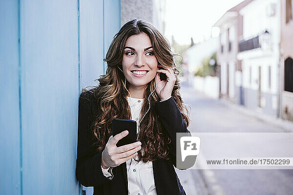 Businesswoman listening music on smart phone leaning on blue wall
